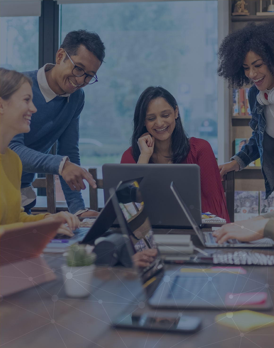 The image shows five associates working inside an office next to each other and interacting with each other. Two of the associates are standing while the other three are sitting down. 